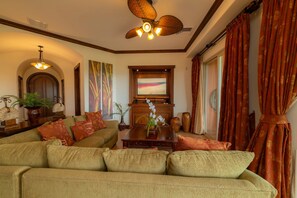 A living room with a sofa, a coffee table, and a television. There is a ceiling fan, a fireplace, and a window with curtains. - Montebello 1E 3 bed 3 bath, ocean view (Playa Herradura)