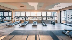 Et rummeligt fitnesscenter med flere løbebånd, crosstrainere og en motionscykel, store vinduer giver udsigt over byen.