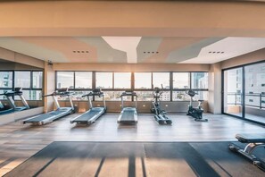 Ein geräumiges Fitnessstudio mit mehreren Laufbändern, Crosstrainern und einem stationären Fahrrad, große Fenster mit Blick auf die Stadt.