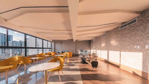 Et moderne loungeområde med runde borde og gule stole, store vinduer der giver udsigt over byen, samt et mønstret gulv.