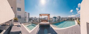 Ein Poolbereich auf dem Dach mit einer Liege und Lounge-Stühlen, umgeben von Stadtgebäuden.