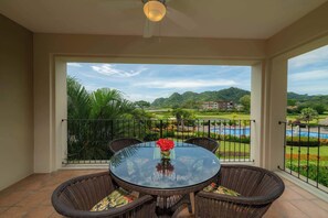 A round glass table with wicker chairs and a vase of red flowers on a balcony with a view of a swimming pool and greenery. - Del Mar 3i - 3 bdr 2 bath Ocean View (Playa Herradura)
