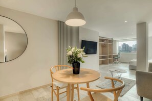 Un salon moderne avec une table ronde en bois, un vase de fleurs, un téléviseur et une vue sur la ville, visible à travers une grande fenêtre.