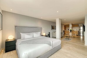 Une chambre d’hôtel moderne avec un grand lit, des tables de chevet, un bureau et, au fond, une petite cuisine.