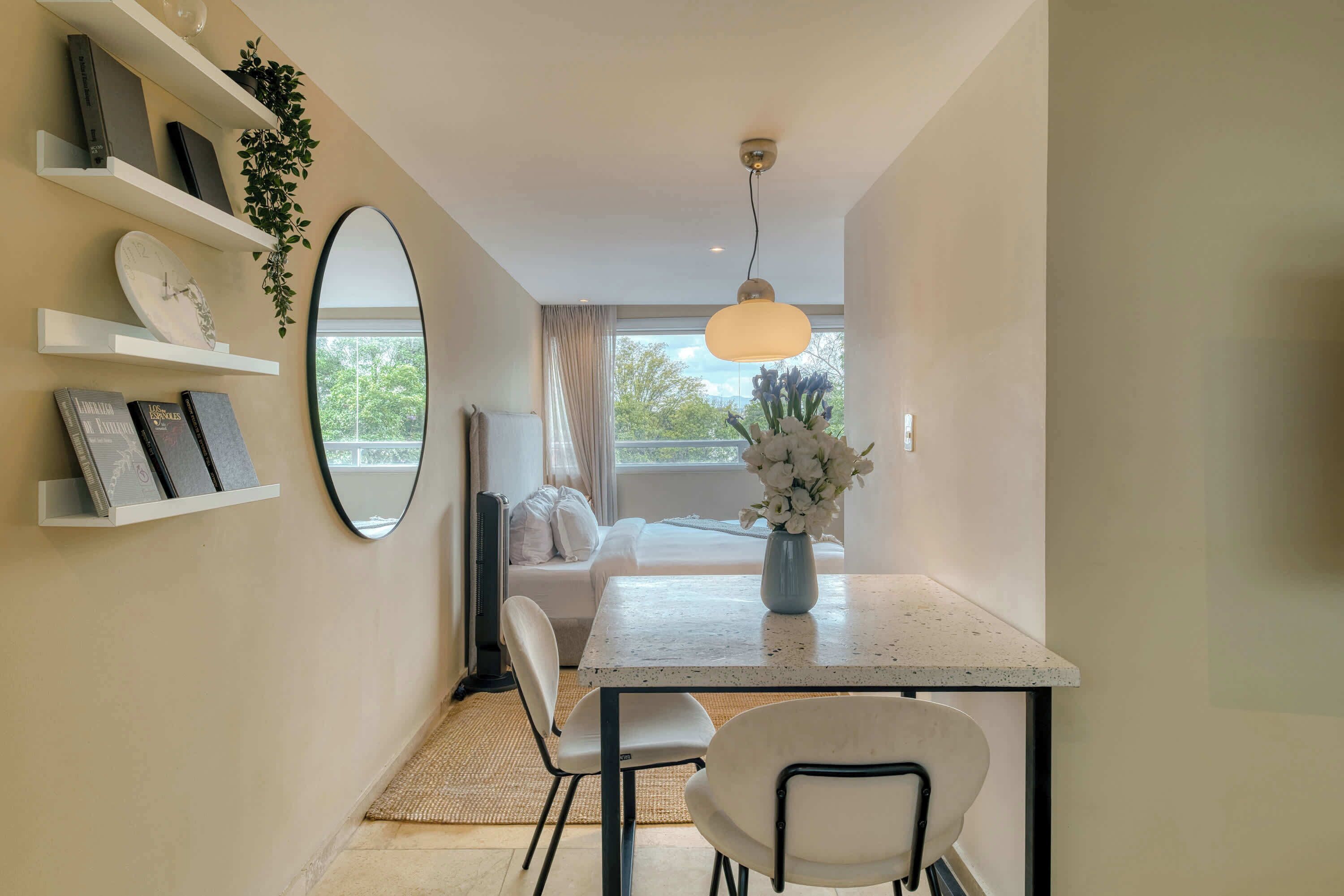 A modern room with a dining table, chairs, a mirror, shelves with books and plants, a bed with white bedding, and a view of the outdoors through a window.