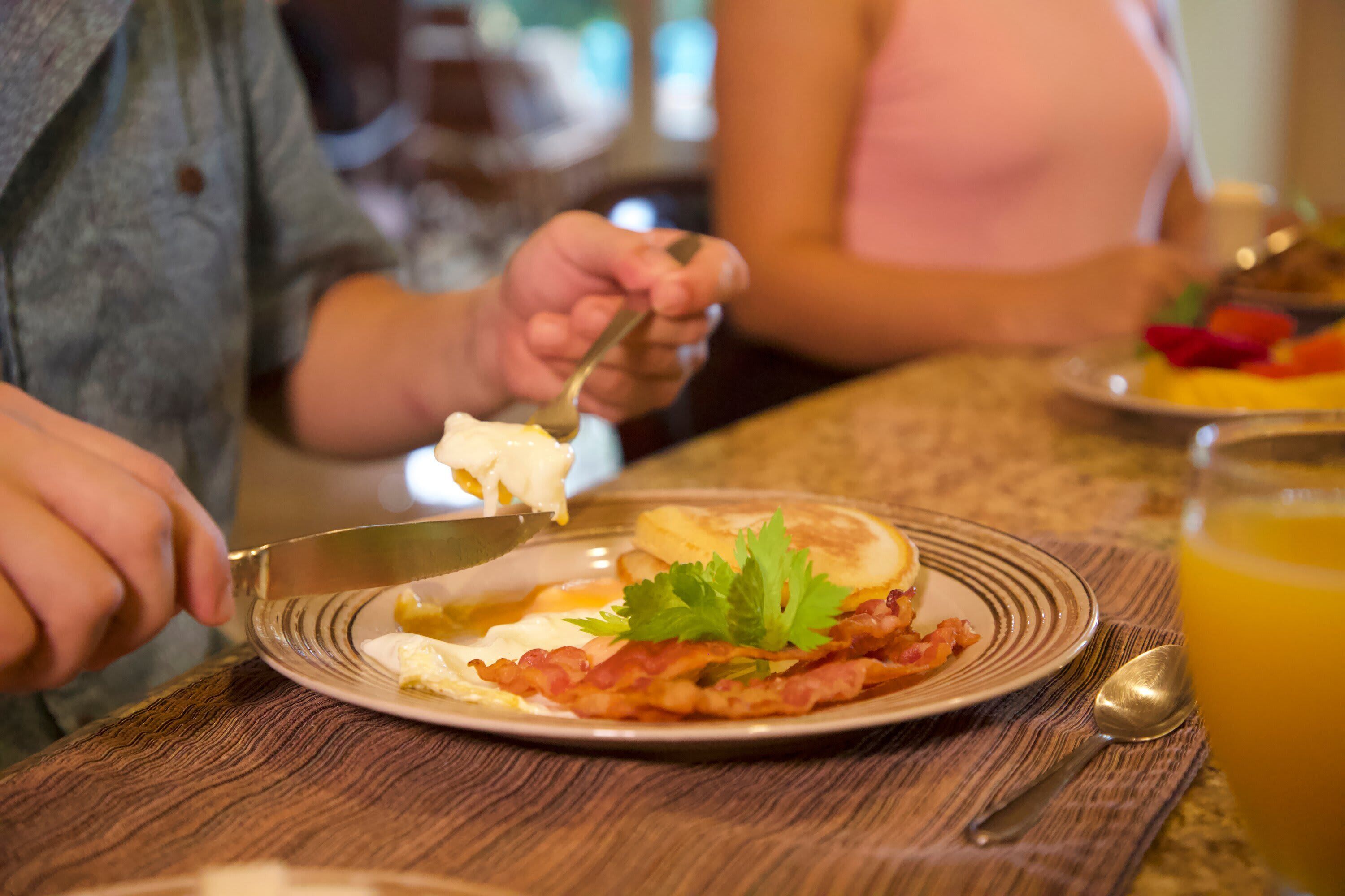 Une table dressée pour le petit-déjeuner avec une assiette contenant des œufs, du bacon et une garniture, un verre de jus d’orange et une cuillère posée sur une table en bois.