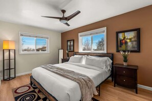 A bedroom with a bed, a ceiling fan, two windows, a nightstand, a lamp, and a painting. - Family Friendly Retreat with Game Room Huge Yard (Arvada)