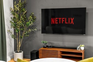 Ein modernes Wohnzimmer mit einem großen Flachbildfernseher an einer grauen Wand, einer Holz-Unterhaltungseinheit darunter und einer Zimmerpflanze links.