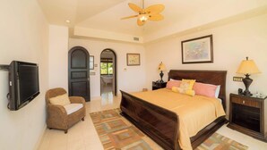 A bedroom with a bed, a television, a chair, a ceiling fan, and a bathroom visible through an archway. - Casa Puesta del Sol 5 bdr - Ocean View Family (Playa Herradura)