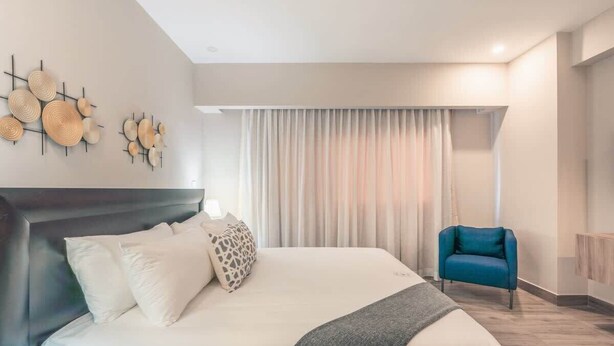 Ett modernt hotellrum med en stor säng, en blå fåtölj och en konstinstallation på väggen.