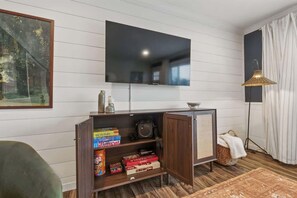 A living room with a flat-screen TV mounted on the wall, a wooden entertainment unit with board games, a floor lamp, and a window with curtains. - Projector Nights & Park Delights I Designer Reno (Arvada)