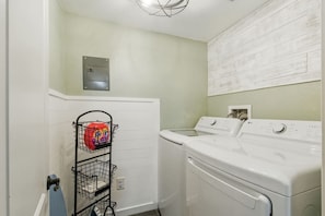 A laundry room with a white washer and dryer, a metal rack with laundry detergent, and a light switch on the wall. - Projector Nights & Park Delights I Designer Reno (Arvada)
