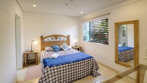 Een slaapkamer met een bed, nachtkastjes, een spiegel, een raam met jaloezieën en uitzicht op een balkon.