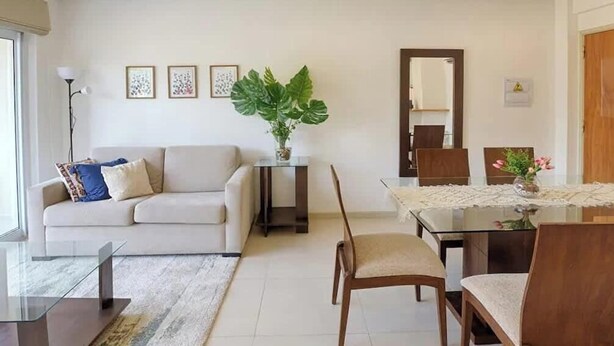 Een woonkamer met een bank, een salontafel en een eettafel met stoelen. Er staat een plant op de salontafel en een spiegel aan de muur.