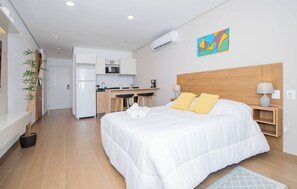 Ein Hotelzimmer mit Bett, Nachttischlampen, einem Schreibtisch mit Stuhl, einer Küchenzeile und einer Wand-Klimaanlage.