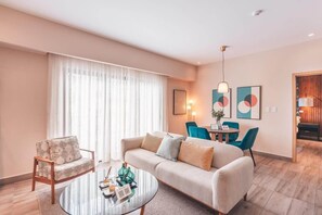 A modern living room with a sofa, armchair, and coffee table. There is a dining area with chairs and a table. The room has large windows with curtains. - Santo Domingo Gem Modern Apartment with Amenities! (Santo Domingo)