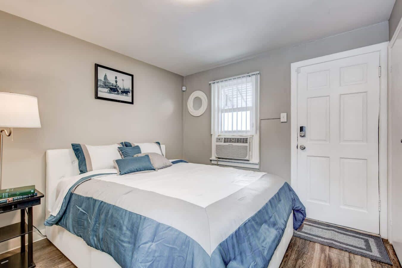 Une chambre à coucher comprenant un lit, une table de chevet, une lampe, un tableau, une fenêtre, un appareil de climatisation et une porte.