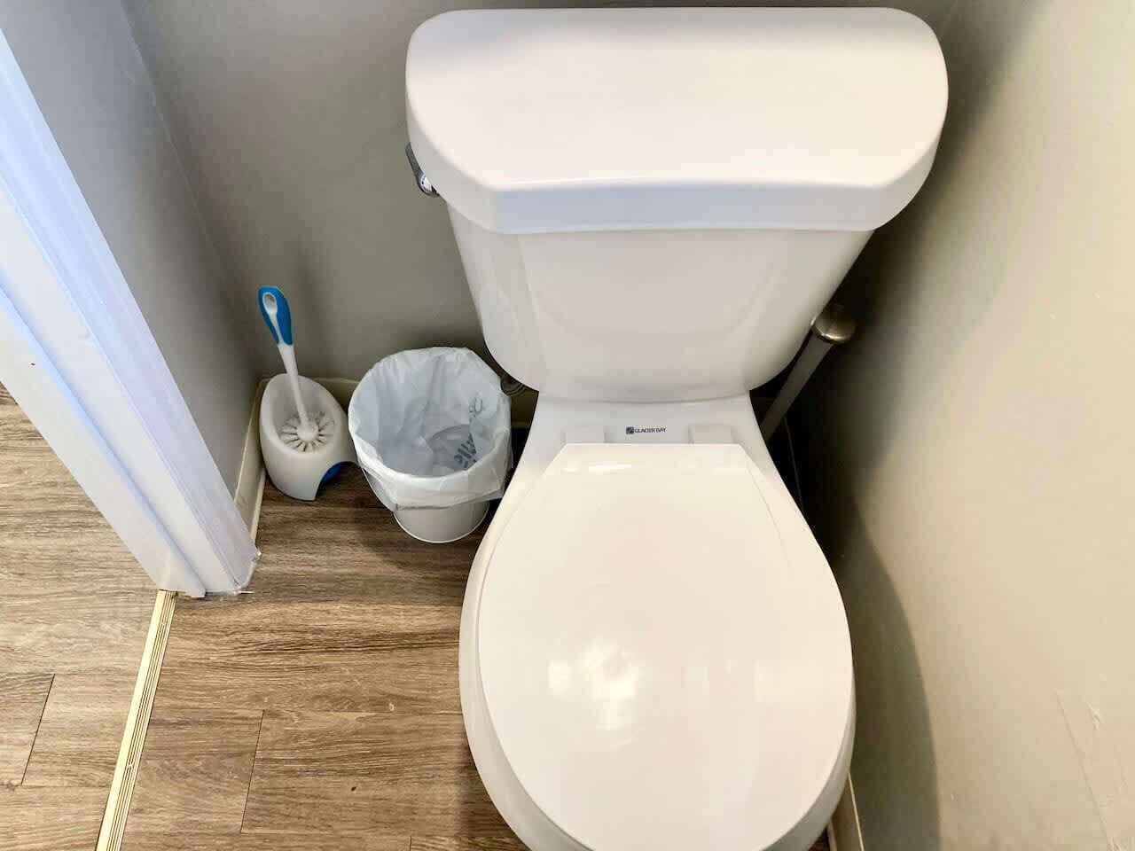 Des toilettes blanches avec un abattant fermé, une poubelle blanche et une brosse à manche bleue dans une salle de bain dont le sol est recouvert de parquet.