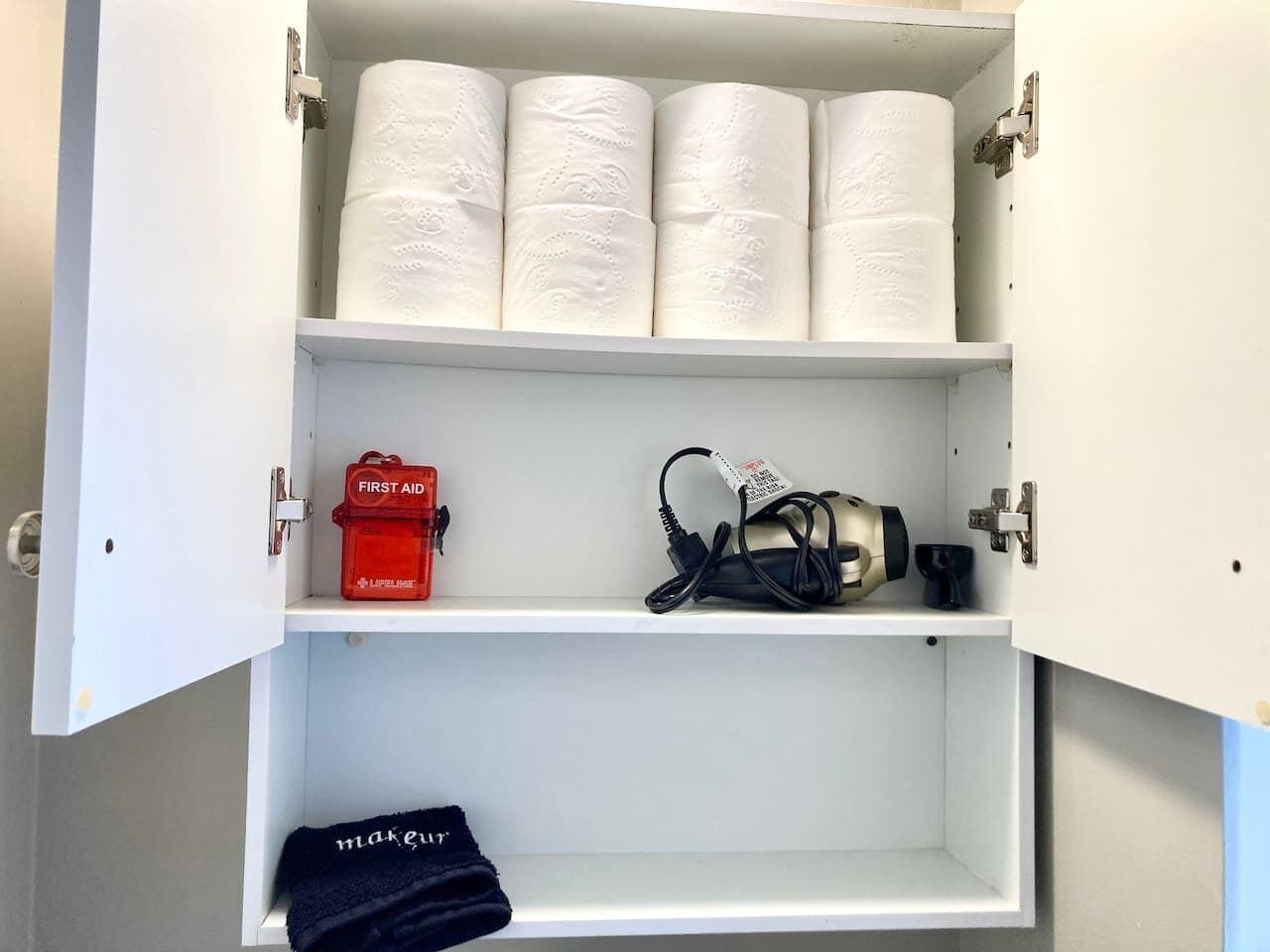 A cabinet with toilet paper, a red first aid kit, and a black electronic device.