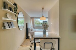 Ein modernes Zimmer mit Esstisch, Stühlen, einem Spiegel, Regalen mit Büchern und Pflanzen, einem Bett mit weißer Bettwäsche und Blick ins Freie durch ein Fenster.