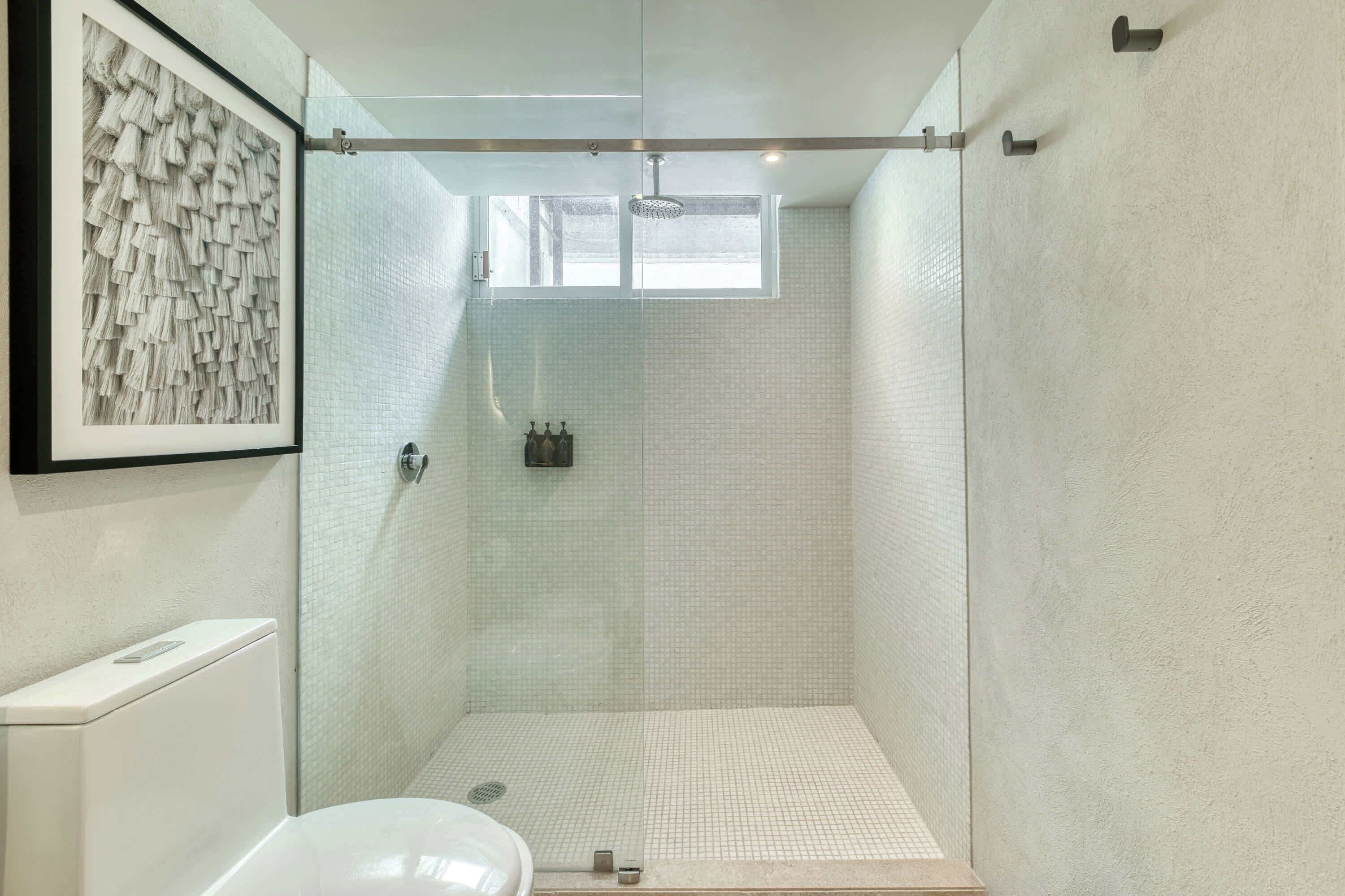 A modern bathroom with a glass shower enclosure, white tiled walls, and a framed artwork on the wall.