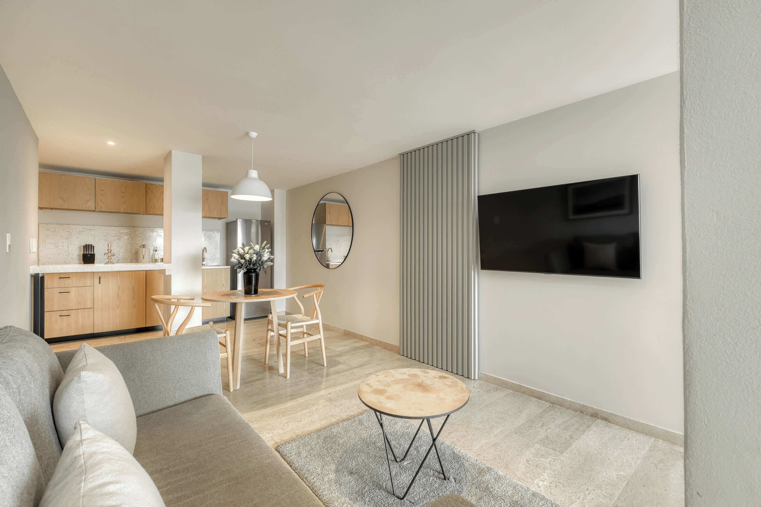 Una sala de estar moderna con un sofá, una mesa redonda de centro, un televisor montado en la pared y una cocina al fondo.