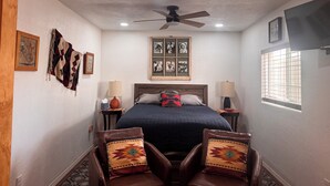A bedroom with a bed, two bedside tables, a ceiling fan, a window with bars, and a TV mounted on the wall. - A GREAT PRICE! FOR A COZY CASITA, SECURED ON A LARGE PRIVATE LOT ALL TO YOURSELF (Yuma)
