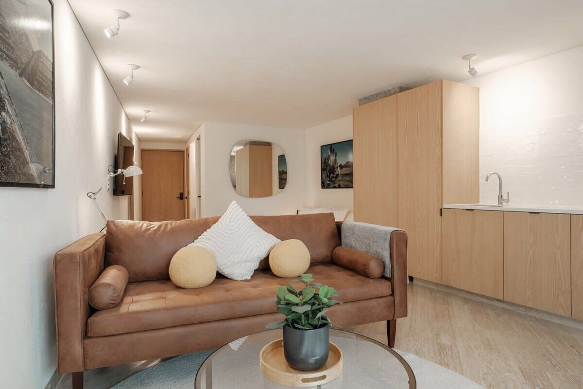 A modern living room with a brown leather sofa, a glass coffee table with a plant, and a kitchen area with wooden cabinets and a white countertop.