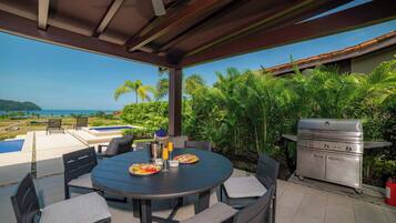 An outdoor patio with a round table, chairs, a grill, and a view of the ocean.