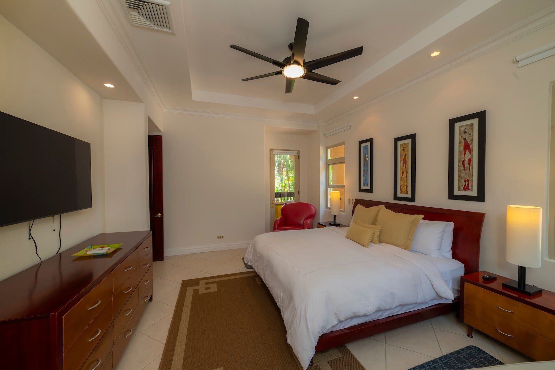 A bedroom with a bed, a TV, a ceiling fan, and artwork on the walls.