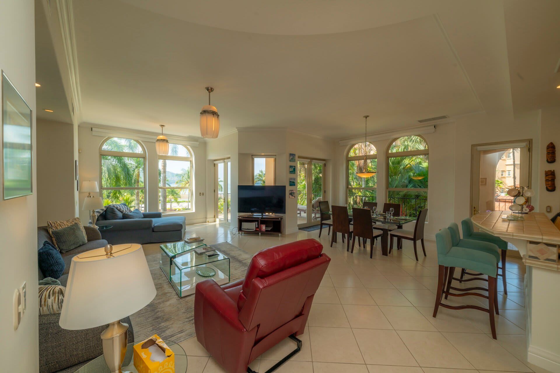 A spacious living area with a sofa, red chair, TV, dining table, and large windows offering a view of palm trees.