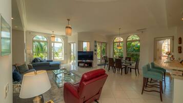 A spacious living area with a sofa, red chair, TV, dining table, and large windows offering a view of palm trees.