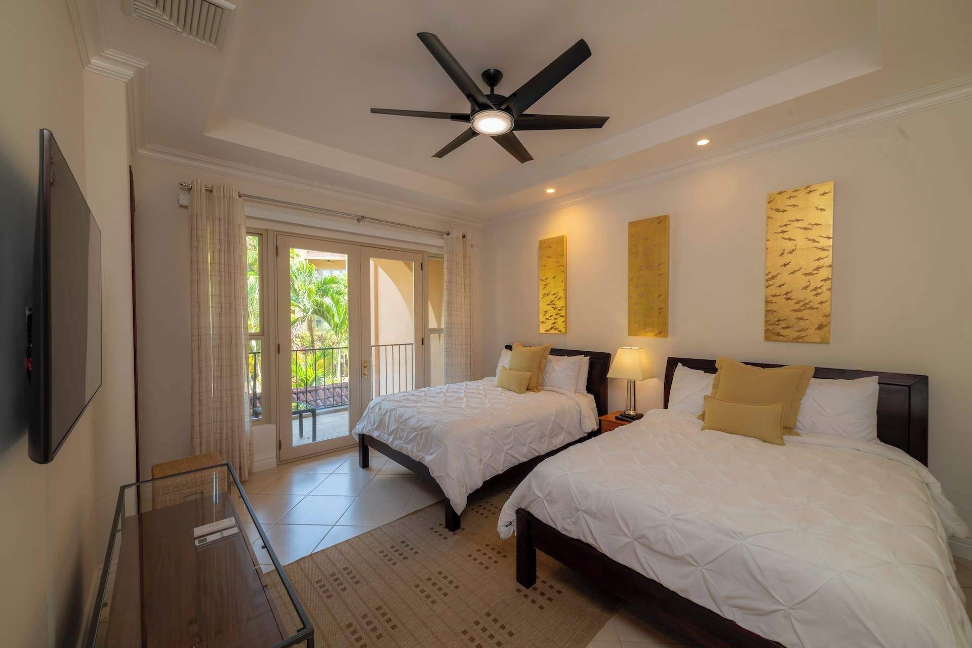 A hotel room with two beds, a ceiling fan, a TV, and artwork on the walls.