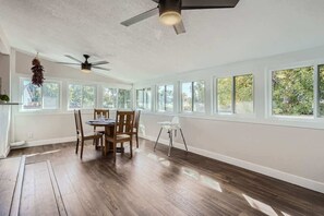 Quarto com mesa de jantar, cadeiras, ventilador de teto e janelas amplas.