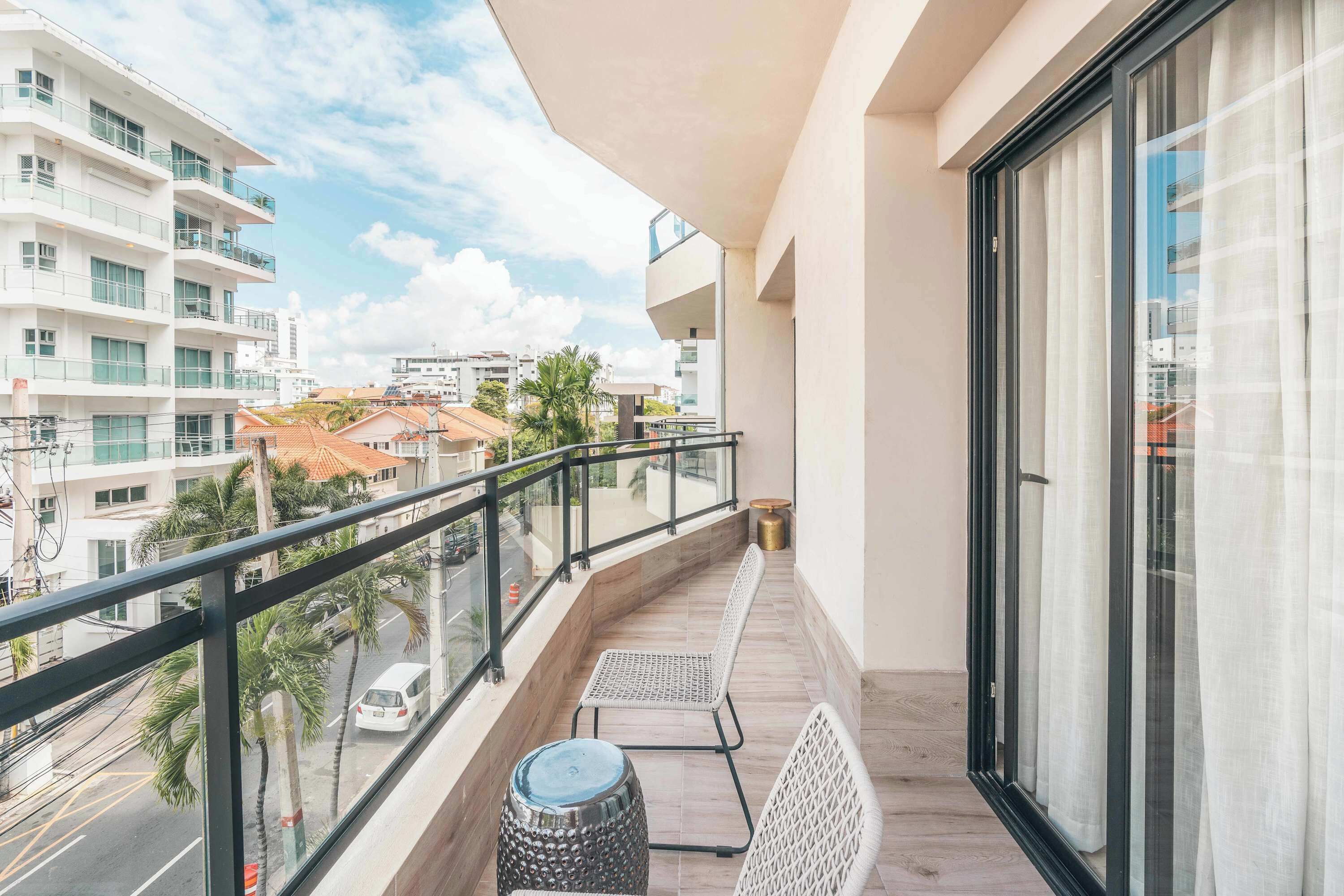 A balcony with a view of the street, a chair, a small table, and a potted plant.