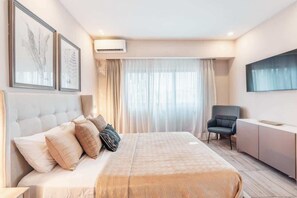 A modern hotel room with a large bed, a desk, and a chair. There is a TV mounted on the wall and an air conditioning unit above the bed. - Stylish Apartment in Piantini Pool & Gym u302 (Santo Domingo)