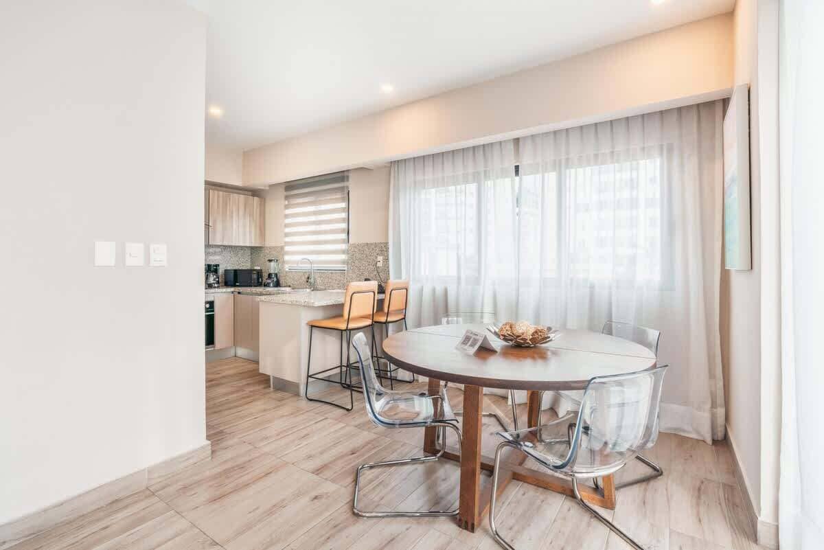 A modern kitchen with wooden cabinets, a dining area with a round table and chairs, and a large window with curtains.