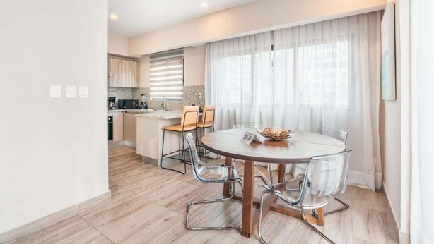 A modern kitchen with wooden cabinets, a dining area with a round table and chairs, and a large window with curtains. - Stylish Apartment in Piantini Pool & Gym u302 (Santo Domingo)