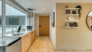 A modern kitchen with a dishwasher, double sink, and wooden cabinets. There is a hallway with a wooden door and a wall-mounted shelf with books and a clock. - Modern King Studio in Central Roma Norte (Ciudad de México)