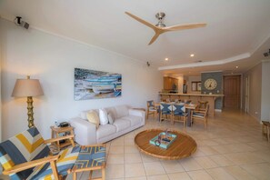 Un salon avec un canapé, des fauteuils, une table basse, un ventilateur de plafond, un tableau et, au fond, une cuisine.