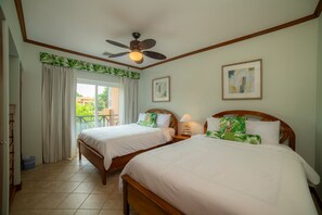 Quarto de hotel com duas camas, um ventilador de teto, uma janela com cortinas e obras de arte nas paredes.