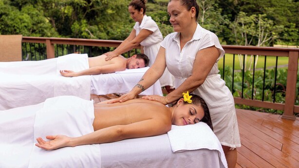Ein Bereich für Massagetherapie mit drei Klienten, die auf einer Holzterrasse Behandlungen erhalten, mit Blick auf üppiges Grün.