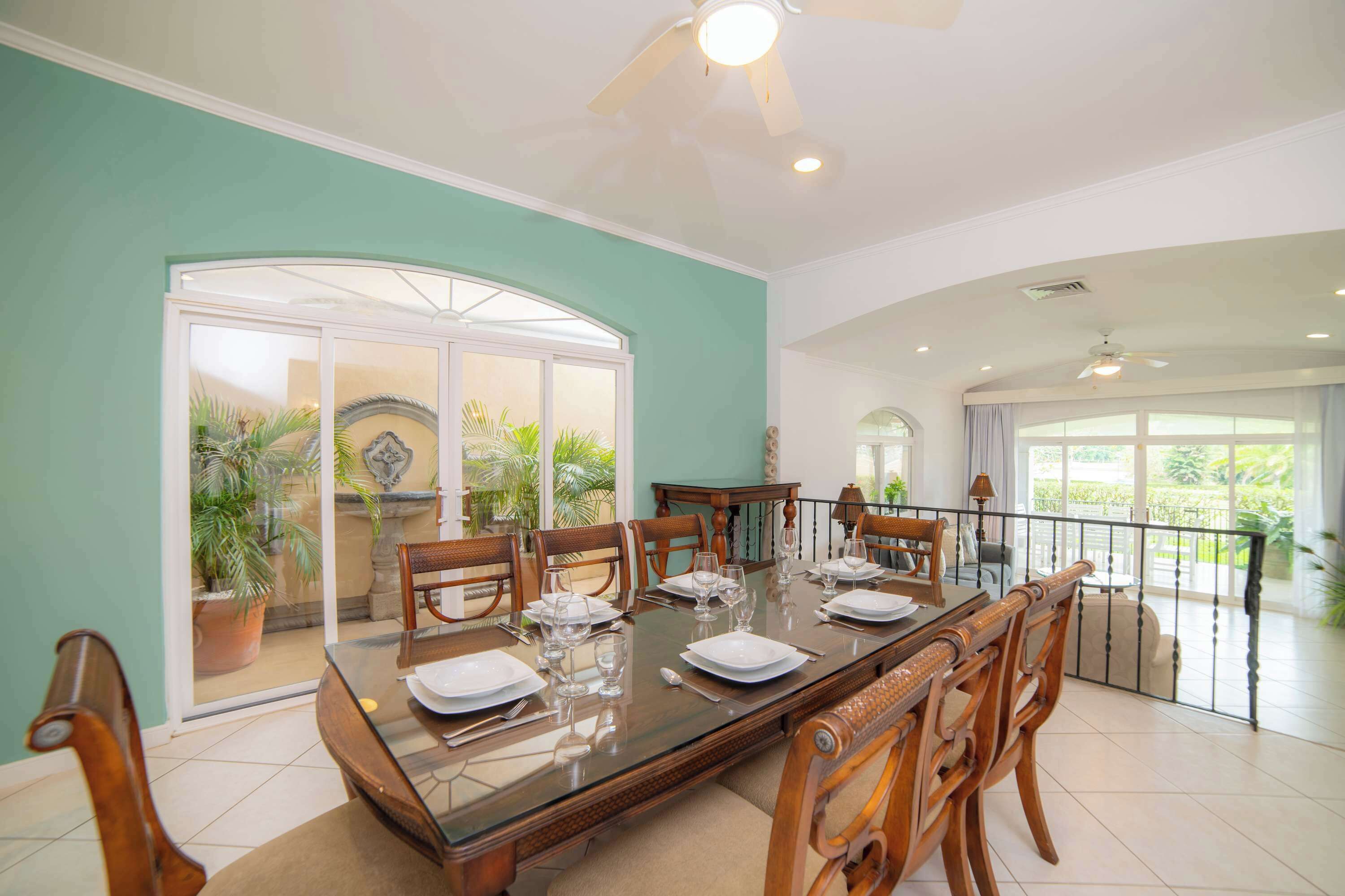 A dining area with a glass table, wooden chairs, and a view of greenery through a glass door.