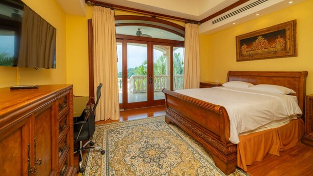 A bedroom with a large bed, a TV, a desk with drawers, a chair, a patterned rug, and a balcony with curtains. - Casa Garmus - 10 bdr Ocean View (Playa Herradura)
