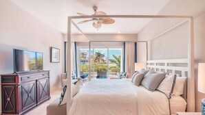 A modern hotel room with a large bed, a ceiling fan, a TV, and a balcony with a view of palm trees. - Apt with 2br-beachfront private pool in Sotogrande (Punta Cana)