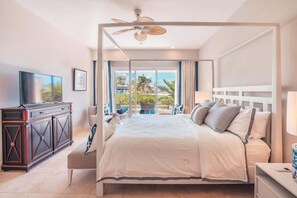 A modern hotel room with a large bed, a ceiling fan, a TV, and a balcony with a view of palm trees. - Apt with 2br-beachfront private pool in Sotogrande (Punta Cana)