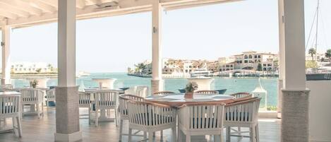 A covered outdoor dining area with white chairs and tables, overlooking a marina and buildings.