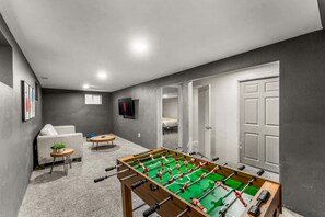 Ein Zimmer mit einem Kicker, einer Couch, einem kleinen runden Tisch mit einer Pflanze und einem an der Wand montierten Fernseher.