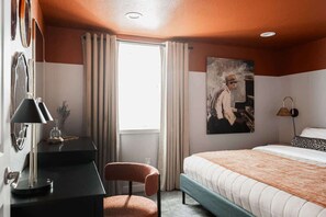 Ein Hotelzimmer mit einem Bett, einem Schreibtisch mit Lampe, einem Stuhl, einem Fenster mit Vorhängen und einem Bild an der Wand.