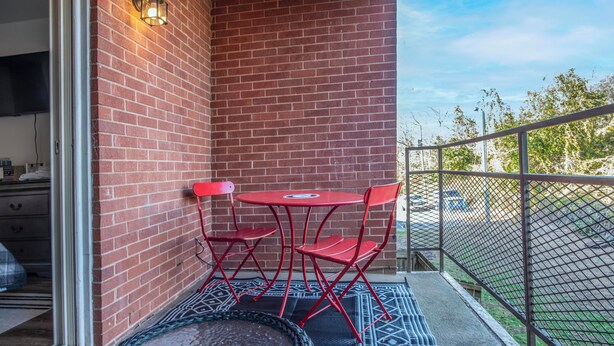 Un balcón con una mesa y sillas rojas, una silla de mimbre y un muro de ladrillo.